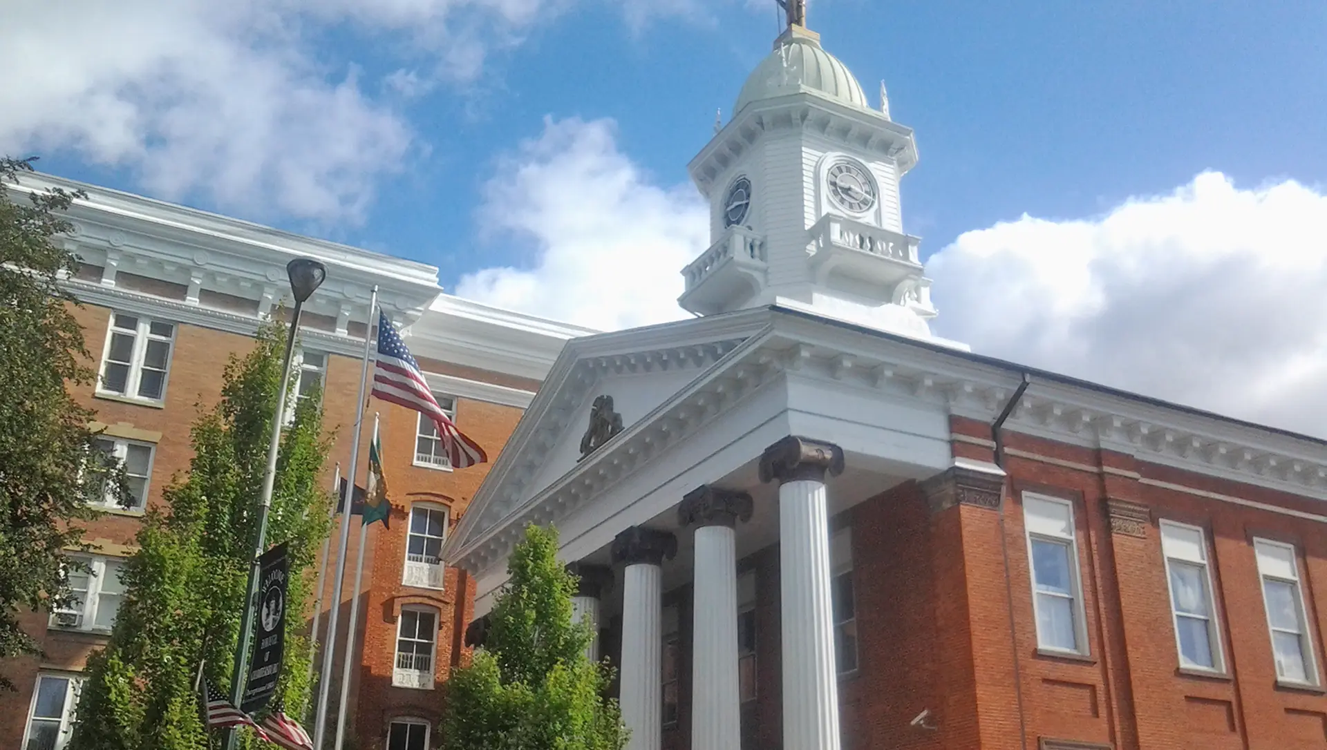 Red-brick government building with white columns and a clock tower, flying American and state flags, under a partly cloudy sky.